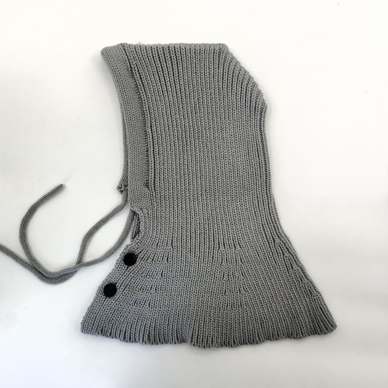 Grå ribbstickad balaklava-huva med justerbara snören och svarta knappar mot vit bakgrund.