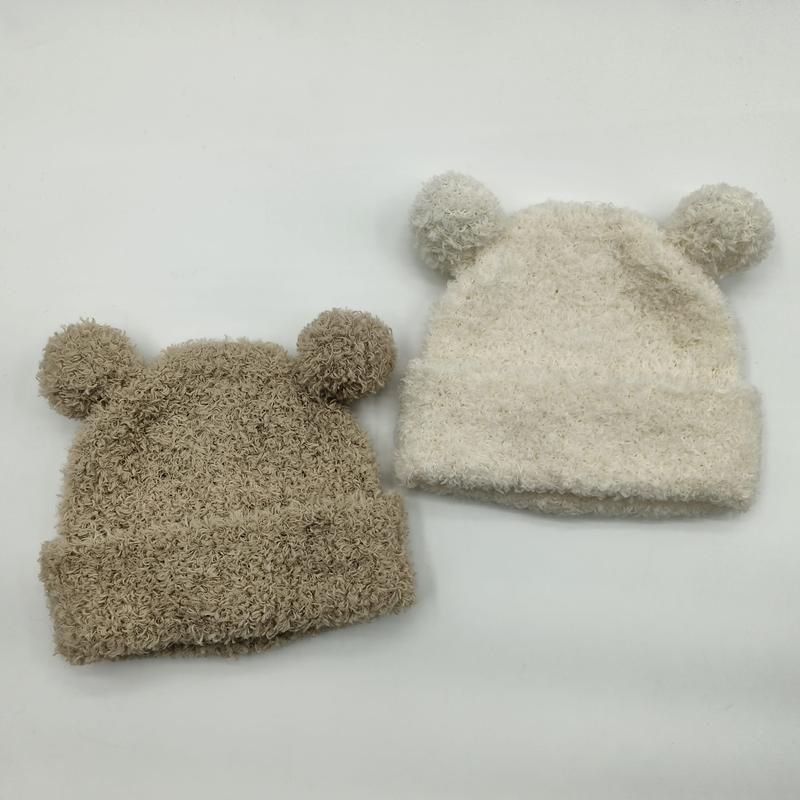 Två stickade babylmössor med björnöron och pompomer i beige och grädde på vit bakgrund.