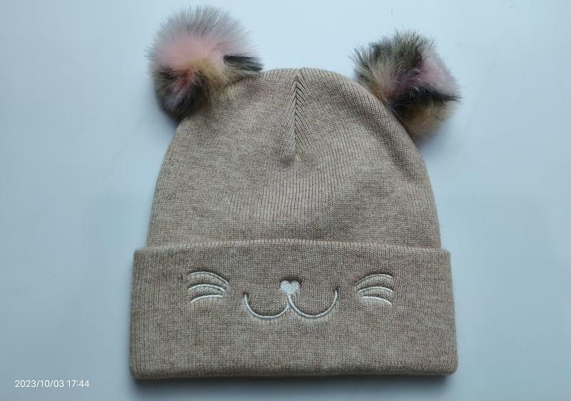 Beige stickad barnmössa med kattansikte och flerfärgade pom poms – grossistleverantör av barnkläder.