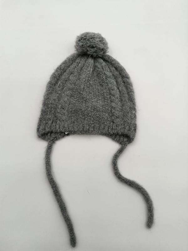 ""Grå stickad babymössa med pompom och knytband på vit bakgrund.""