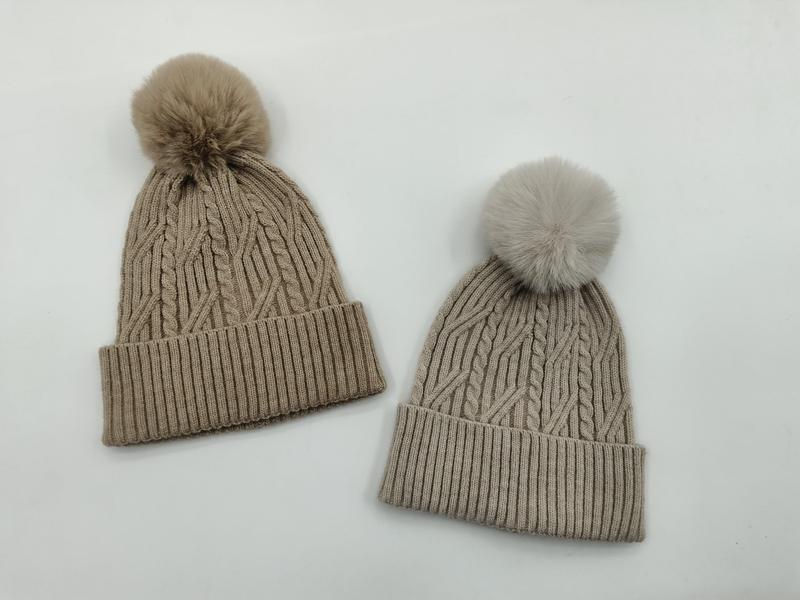 Två kabelstickade vintermössor med fluffiga pom poms i taupe och ljusgrå mot vit bakgrund.