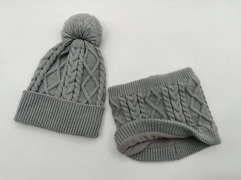 Grå kabelstickad mössa med pom pom och matchande fleecefodrad halsvärmande set mot vit bakgrund.