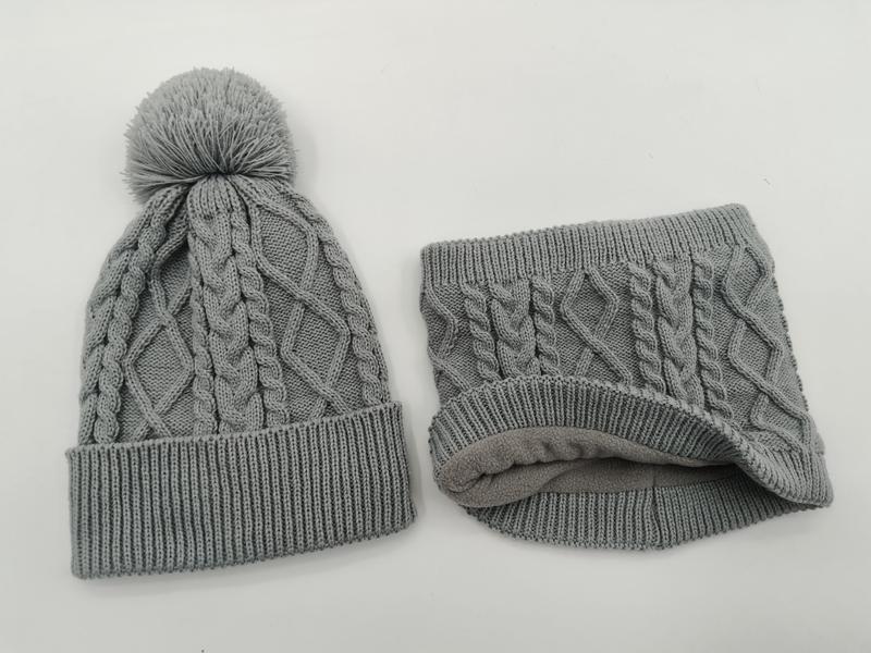 Grå kabelstickad vintermössa med pompom och matchande fleecefodrad halsvärmarsats på vit bakgrund.