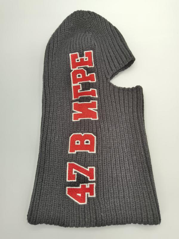 Grå ribbstickad balaclava med röd och vit broderad text på mörkgrå ullfärgat tyg.