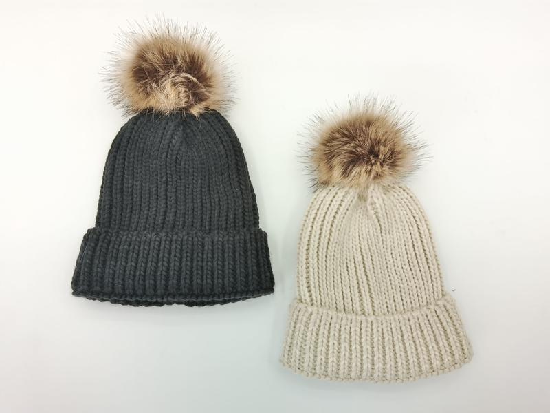 Två grova stickade mössor med fejkpälspom-pom i mörkgrå och cremevit mot vit bakgrund.