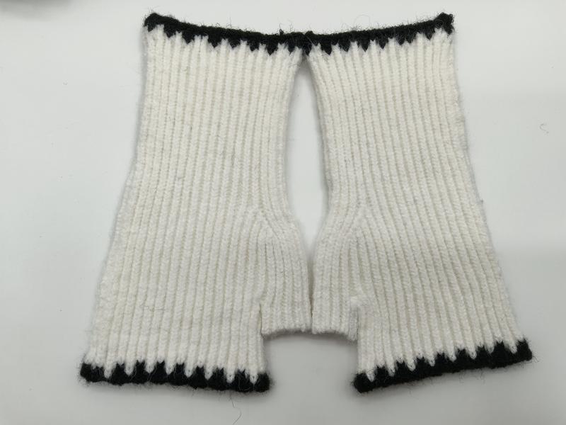 Handstickade vita fingerlösa vantar i ribbat mönster med svarta detaljer vid muddar och handledsöppningar.