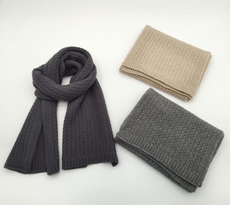 Tre kabelstickade ullhalsdukar i antracit, ljusgrå och beige – en draperad, två vikta, mot vit bakgrund.