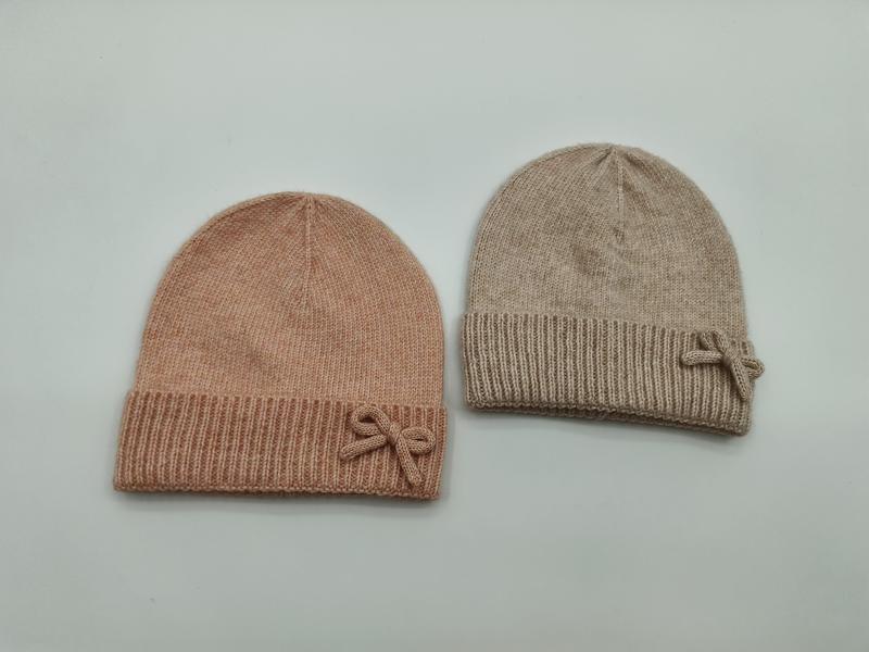 Två mjuka stickade mössor med rosettdetalj, en i blush rosa och en i beige, med ribbad kant. Set om 2.