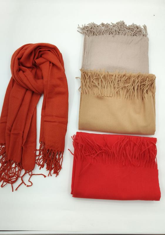 Fyra pashminasjalar med fransar i roströd, beige, kamel och röd ton, en draperad och tre vikta på vit yta.
